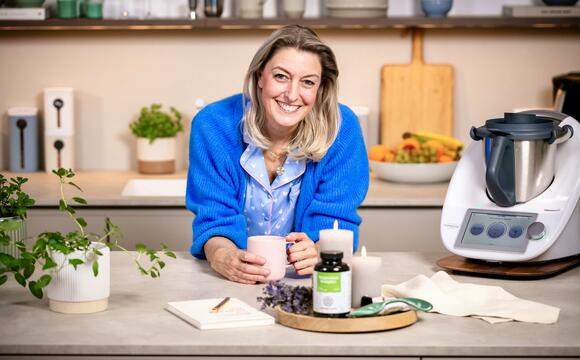Blonde Frau in einer Küche, auf die Arbeitsplatte gestützt mit einem Becher in beiden Händen, daneben ein Thermomix®