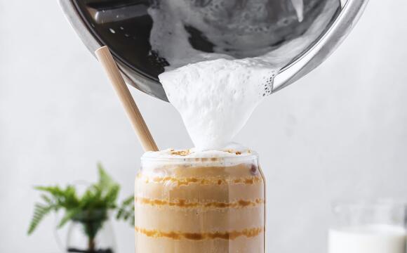 Ein Glas mit Milchkaffee, jemand gießt Milchschaum mit dem Thermomix® darauf