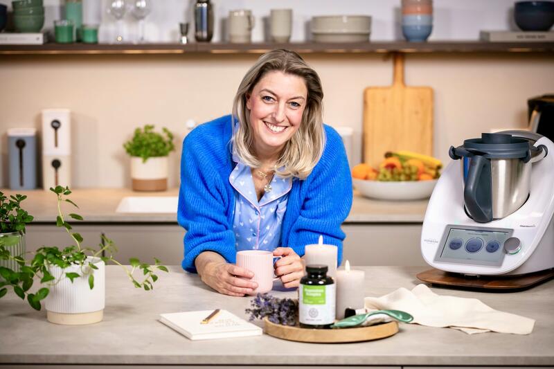 Blonde Frau in einer Küche, auf die Arbeitsplatte gestützt mit einem Becher in beiden Händen, daneben ein Thermomix®