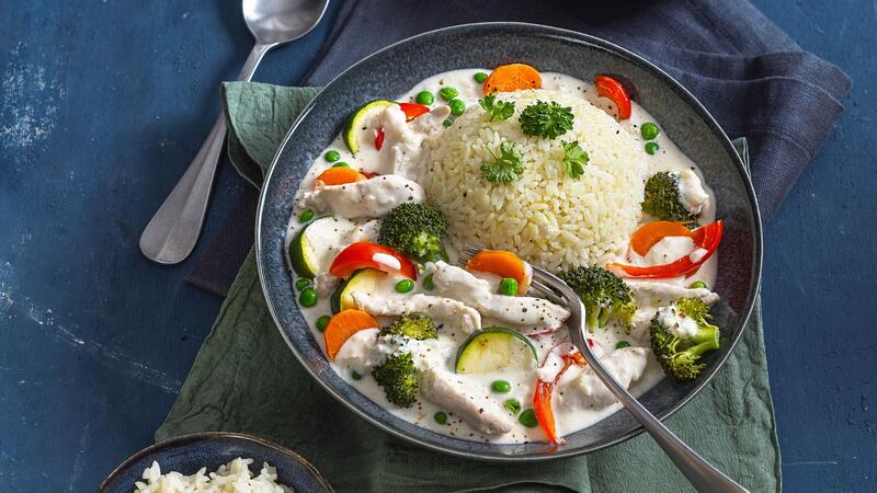 Ein Teller mit Hähnchen-Geschnetzeltem in heller Soße, dazu Reis, Paprika, Brokkoli, Blauer Untergrund