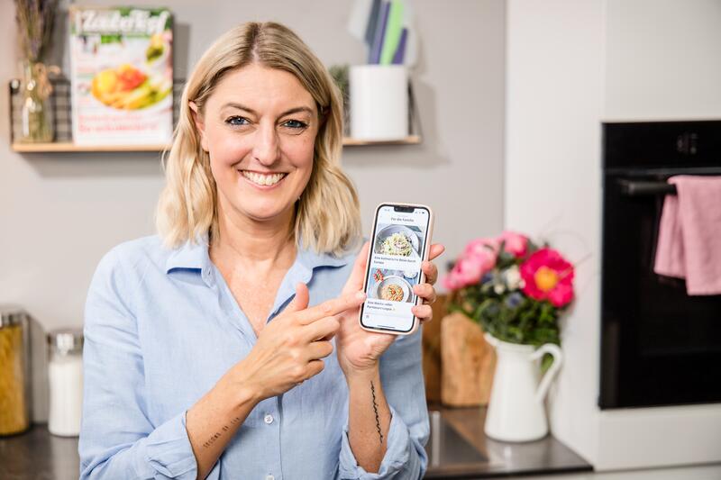 Desi hält ein Smartphone mit der ZauberTopf App hoch