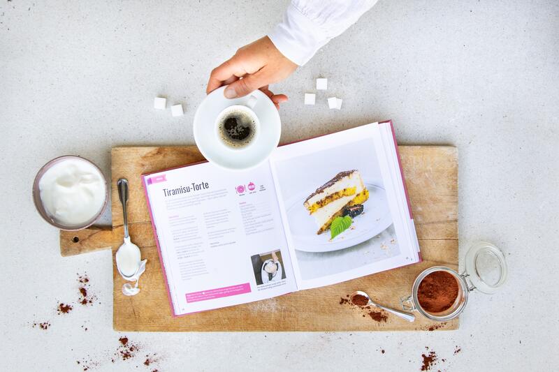Das Kochbuch Südtirol backt! liegt aufgeschlagen mit dem Rezept für eine Tiramisu-Torte auf einem Brett. Eine Hand reicht einen Espresso ins Bild.