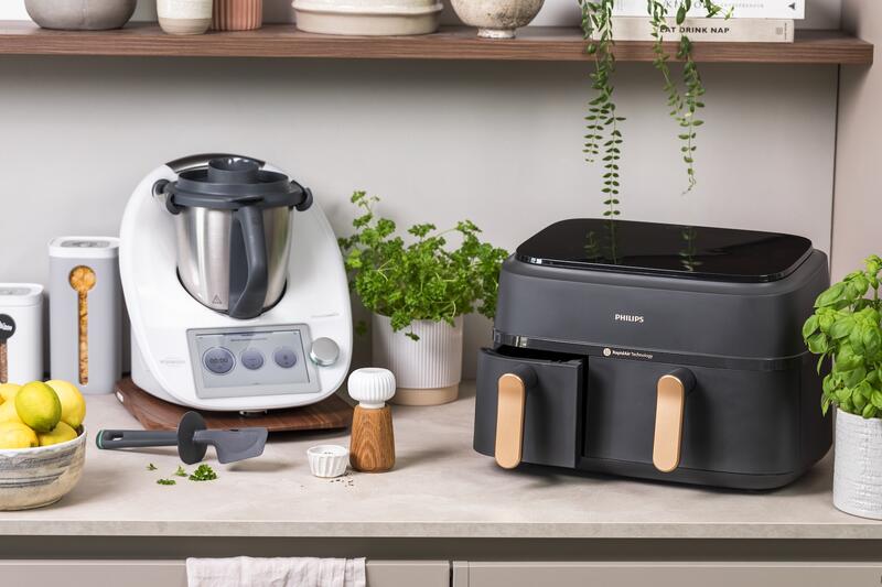 Thermomix® und Airfryer auf einer Arbeitsplatte, daneben Töpfe mit Kräutern