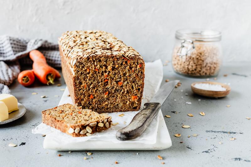 Angeschnittenes Vollkornbrot mit Karotten auf einem hellen Brett, daneben ein Messer