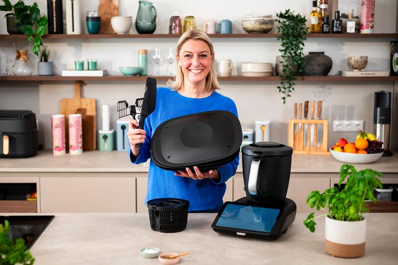 Eine blonde Frau zeigt den Thermomix® TM7 und sein Zubehör