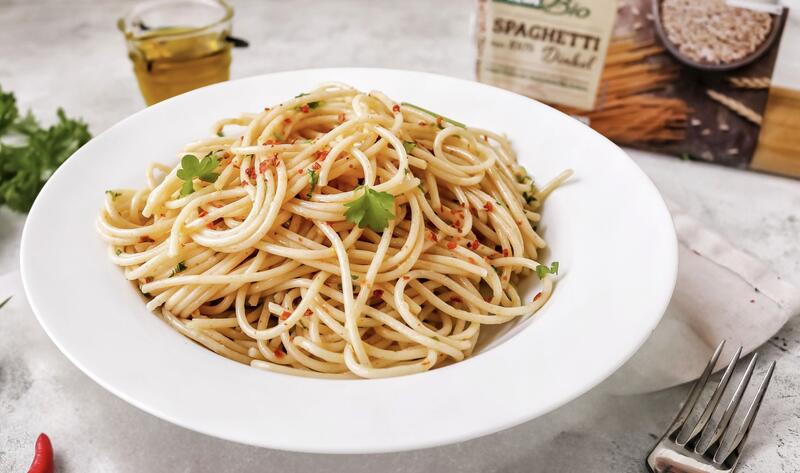 Ein Teller mit Spaghetti auf heller Tischdecke, daneben Peperoni, Besteck und eine Packung Spaghetti