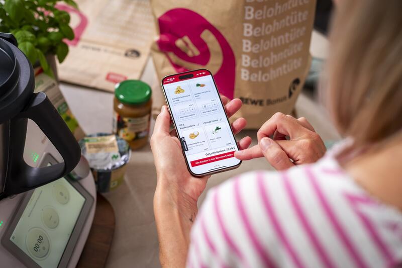 Eine Frau hält ihr Smartphone in der Hand, darauf ist der Einnkaufswagen von REWE online zu sehen