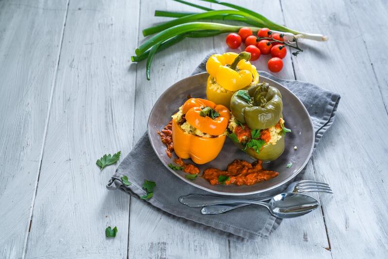 Drei gefüllte Paprika, gelb, orange, grün auf einem Teller, daneben Tomaten und Lauchzwiebeln