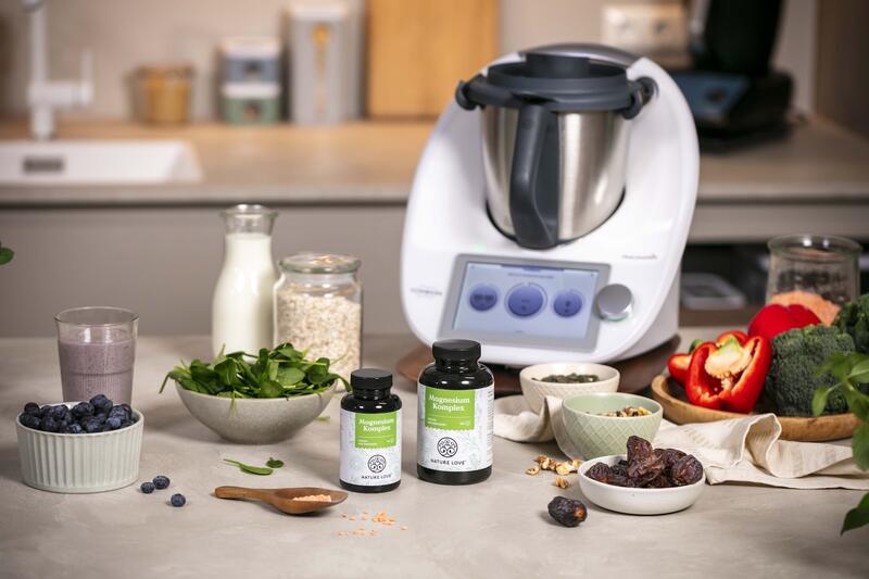 Der Thermomix® umgeben von Gemüse, Dateln und zwei Gläsern mit Magnesium-Tabletten