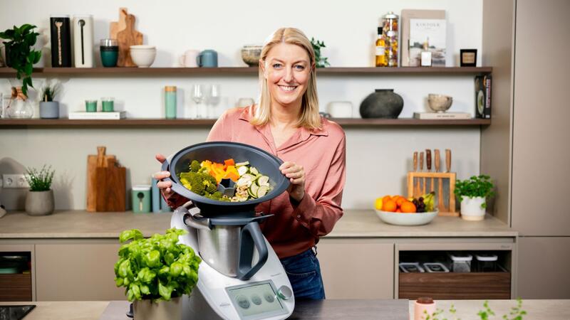 Blonde Frau hält den Varoma® des Thermomix® in beiden Händen, darin ist Gemüse