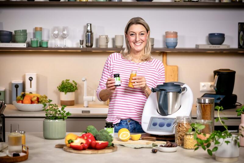 Blonde Frau in gestreiftem Shirt in einer Küche mit Thermomix®. In der Hand ein Saft und ein Glas mit Eisentabletten