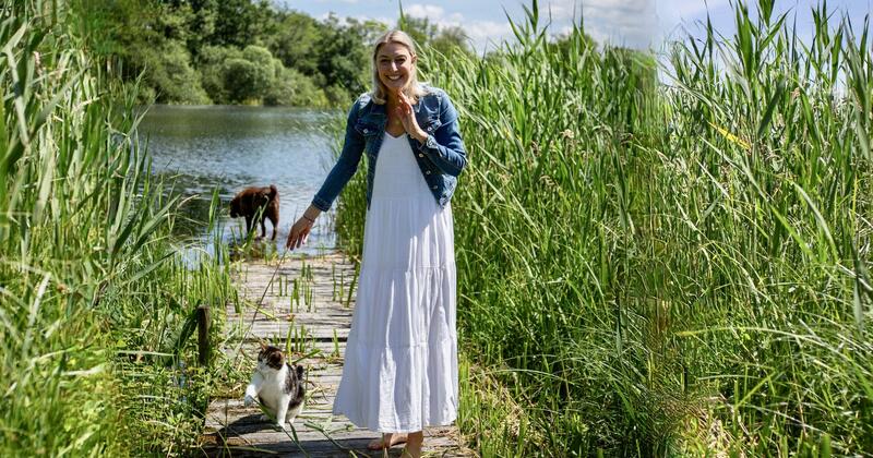 Blonde Frau auf einem Steg im Grünen mit einer Katze, im Hintergrund ein Hund