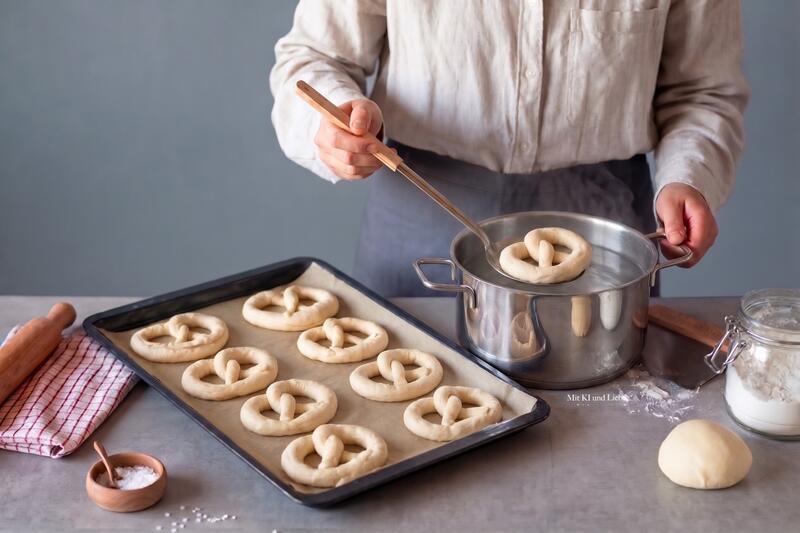Jemand hält eine Kelle mit einer Brezel darauf und gibt sie in einen Topf mit Lauge, daneben ein Blech mit Brezeln