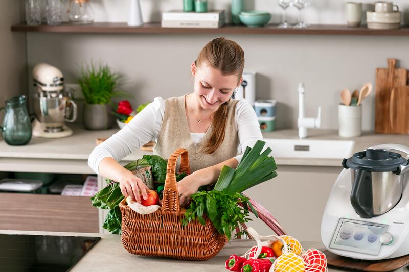 Eine junge Frau schaut in ihren Einkaufskorb voller Gemüse, daneben ein Thermomix®