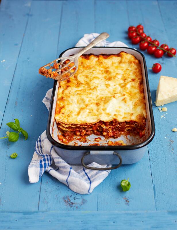 Angeschnittene Lasagne in Auflaufform auf blauem Holztisch.
