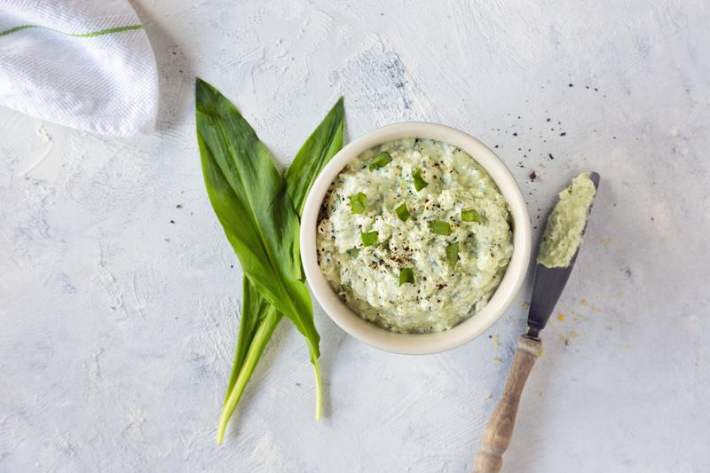 Topshot einer weißen Schale mit Bärlauchfrischkäse aus dem Thermomix® mit zwei angelegten Bärlauchblättern und Messer