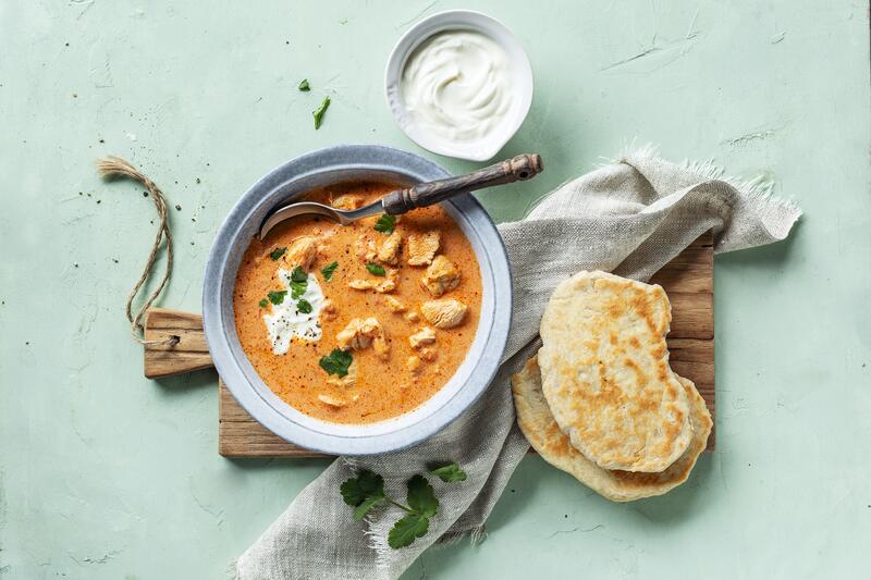 Ein heller Teller mit rotem Hähnchencurry aus dem Thermomix® mit angelegtem Naanbrot auf hellgrünem Untergrund