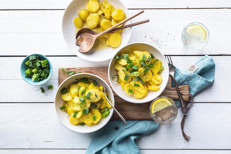 Zwei weiße Teller mit schwäbischem Kartoffelsalat aus dem Thermomix® auf Holzbrett mit angestellten Frühlingszwiebeln