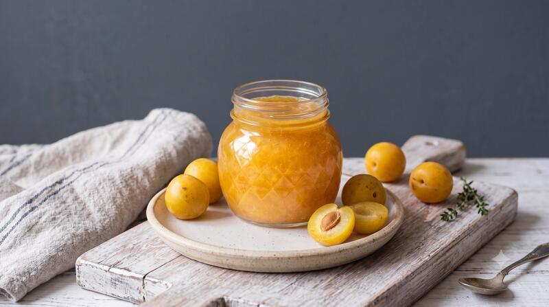 Ein Glas mit gelber Marmelade auf deinem grauen Teller, daneben liegen Mirabellen