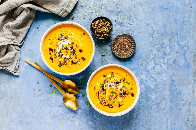 Zwei tiefe weiße Schalen mit Low-Carb-Kürbissuppe aus dem Thermomix® auf blauem Untergrund, mit angelegten goldenen Löffeln, von oben fotografiert
