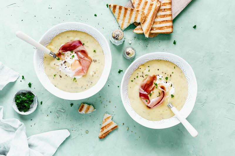 Zwei weiße Schüsseln mit Kartoffelsuppe aus dem Thermomix® mit Räucherschinken auf hellblauem Untergrund