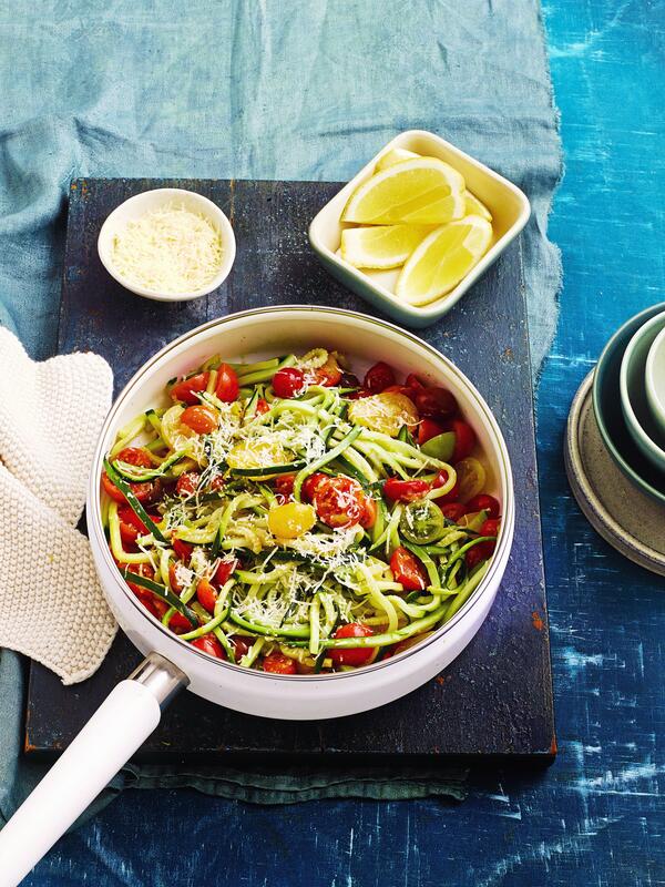 Weiße Pfanne mit Zucchini-Tomaten-Spaghetti auf schwarzem Brett mit angelegten Zitronenspalten