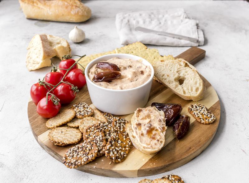 Dattel-Dip in kleiner weißer Schale auf Holzbrett mit Crackern und Tomatenstrauch