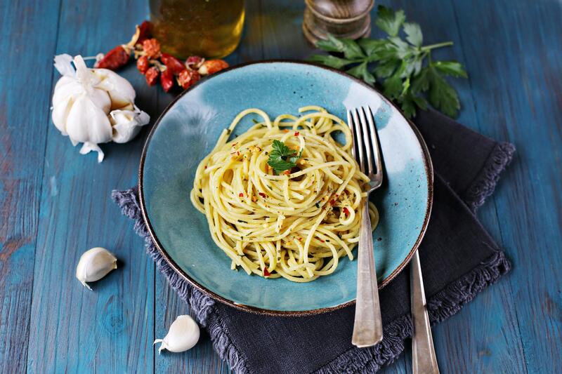 Eine blaue Holzplatte mit einer dunklen Serviette. Darauf ein Teller mit Spaghetti, garniert mit Pfeffer und Basilikum.
