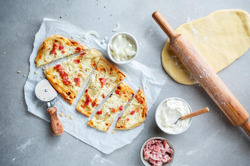 Ausgerollter Flammkuchenteig und Elsässer Flammkuchen auf grauem Untergrund