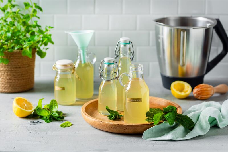 Mehrere Flaschen mit Sirup, im Hintergrund ein Topf mit Minze und der Thermomix®