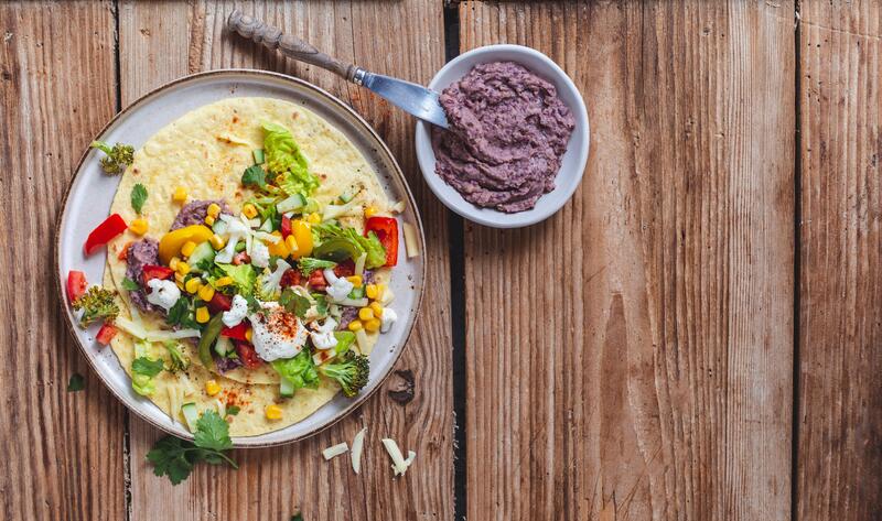 Tortilla mit Gemüse befüllt auf einer Holzplatte, daneben ein Schälchen mit Bohnenmus