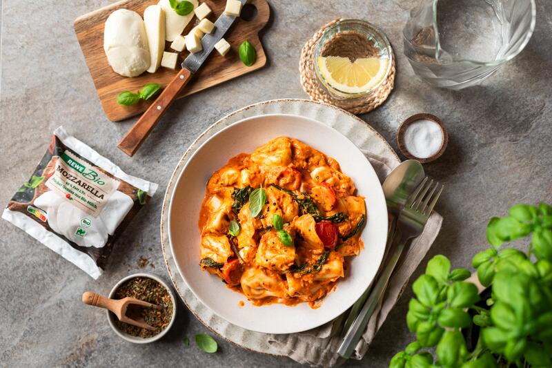 Tiefer Teller mit Tortellini in Tomaten-Mozzarella-Sauce, daneben Mozarella auf einem Brett zerkleinert