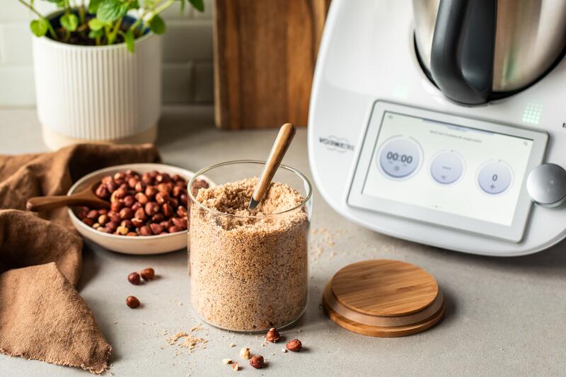 Haselnüsse in einer Schüssel, davor ein Glas mit fertig gemahlenden Haselnüsse, dahinter der Thermomix®