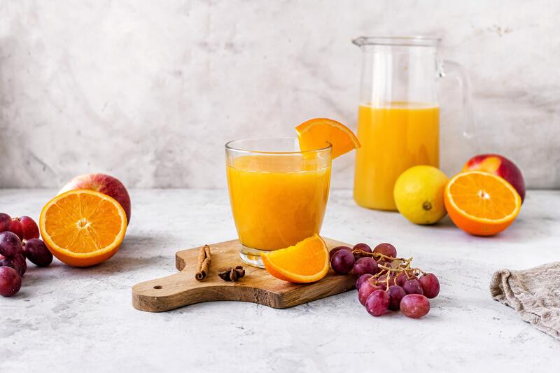 Ein Glas mit orangefarbenem Saft, danaben verschiedene Früchte und eine Karaffe im Hintergrund