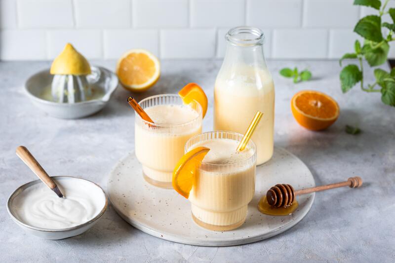 2 Gläser und eine Flasche gefüllt mit leicht gelbem Joghurt-Saft, dekoriert mit Orangen und Zitronen