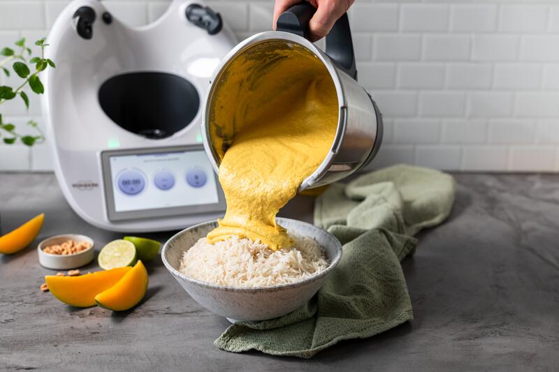 Röstgemüse-Currysoße wird aus dem Mixtopf auf eine Schüssel Reis gekippt, im Hintergrund sieht man den Thermomix® und verschiedenes Gemüse