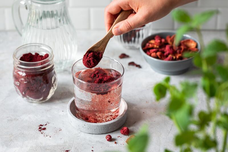 Glas mit Wasser, eine Hand schüttet rotes Immun-Pulver mit einem Holzlöffel hinein