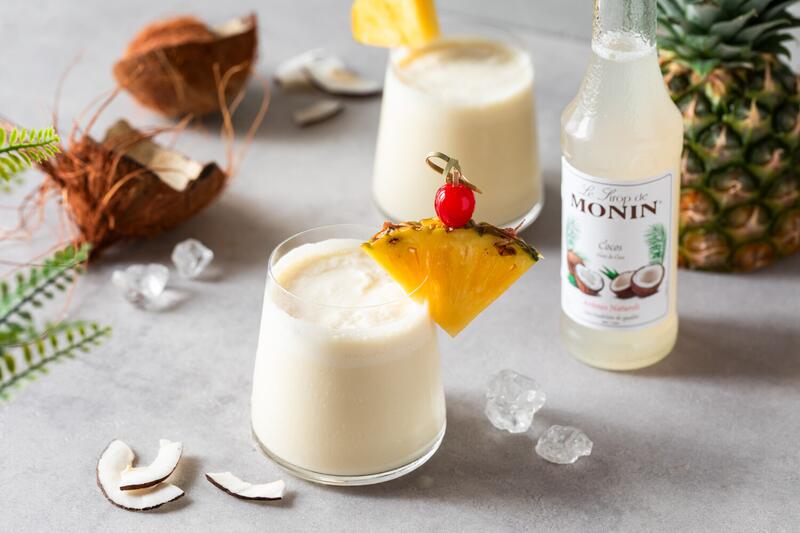 Zwei Gläser Virgin Colada mit einer halbierten Kokosnuss und Ananas im Hintergrund
