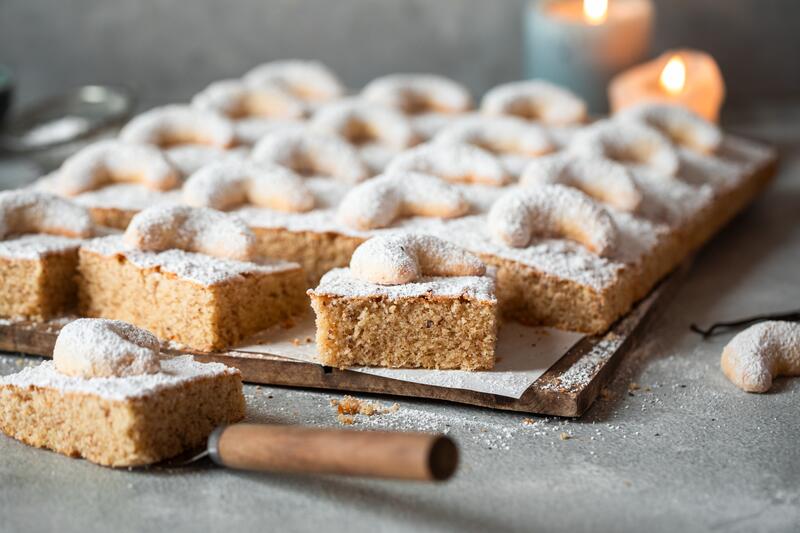 Blech mit angeschnittenem Blechkuchen, auf jedem Stück liegt ein Vanillekipferl und Puderzucker