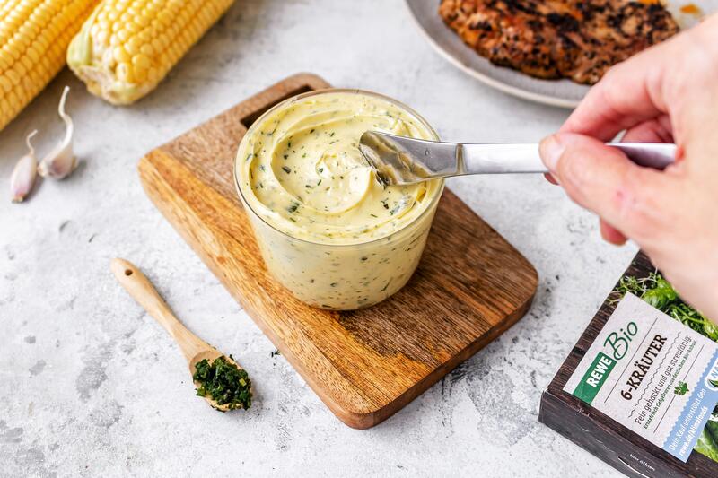 Ein Glas mit Grillbutter, jemand hält ein Messer in der Hand und nimmt etwas davon. Daneben eine Packung 6-Kräuter