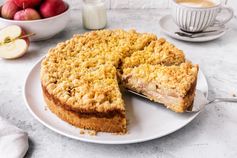 Angeschnittener Apfelkuchen mit Streusel oben drauf, ein Stück wird herausgehoben