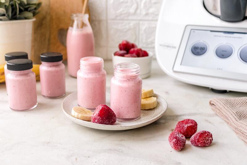 Kleine Gläschen mit rosa Actimel, daneben liegen gefrorene Erdbeeren und Bananen
