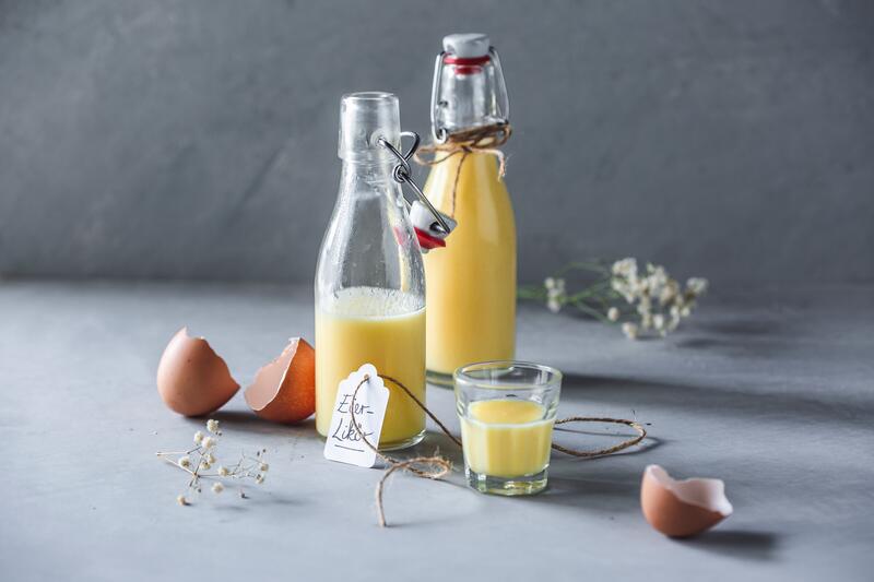 Eierlikör in Flaschen. Im Vordergrund steht ein gefülltes Likörglas.