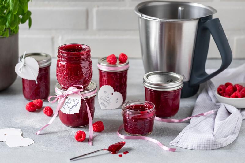 Mehrere Gläser gefüllt mit Himbeermarmelade, Mixtopf im HIntergrund