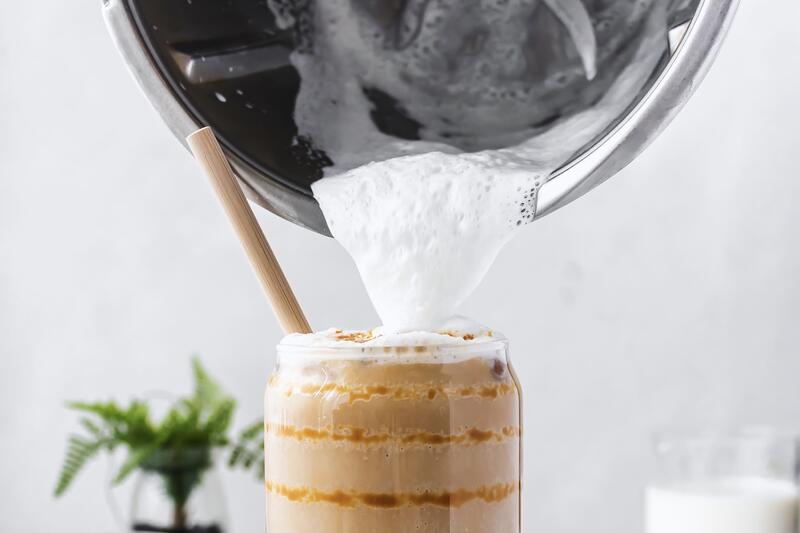 Ein Glas mit Milchkaffee, jemand gießt Milchschaum mit dem Thermomix® darauf
