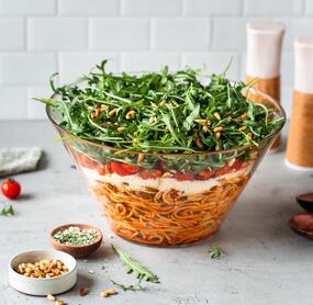 Eine Glasschale mit Schichtsalat. Unten Spaghetti, dann Käse, dann Tomaten, oben Ruccola