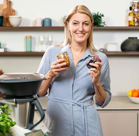 Blonde Frau in einer Küche mit Thermomix®. In der Hand zwei Gläser Marmelade