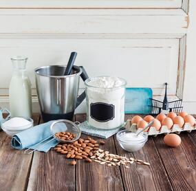 Backzutaten wie Eier, Mehl, Zucker, Mandeln und ein Mixtopf des Thermomix® auf einer Arbeitsplatte