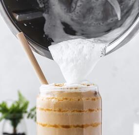 Ein Glas mit Milchkaffee, jemand gießt Milchschaum mit dem Thermomix® darauf