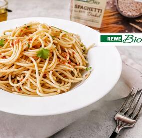 Ein Teller mit Spaghetti auf heller Tischdecke, daneben Peperoni, Besteck und eine Packung Spaghetti